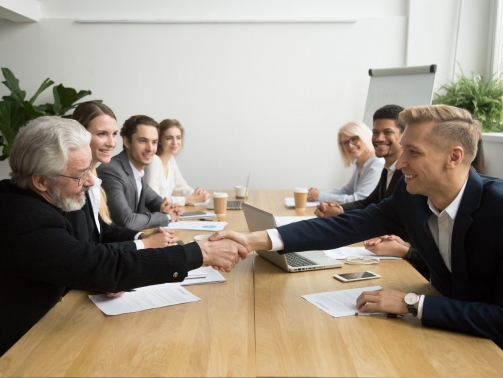 Young and old male partners shaking hands after signing contracts at group multiracial meeting, senior investor buying startup promising support handshaking entrepreneur, partnership deal concept