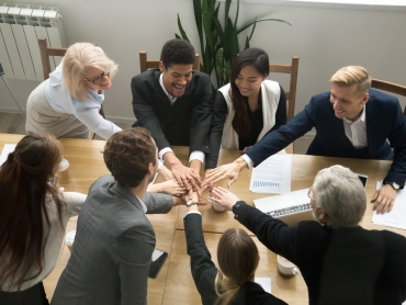 Young and old multiracial business people join pile of stacked hands together at group meeting conference table promising help support, concept of successful teamwork, team building unity, top view